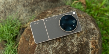 Rear view of the OPPO Find X9 Ultra in Tundra Umber standing vertically on a stone surface with a lush green bokeh background. The device features an eco-friendly vegan leather back with a classic stitching design , vertically oriented Hasselblad and OPPO logos , and a large circular Master Lens camera module with a bronze-toned knurled ring.